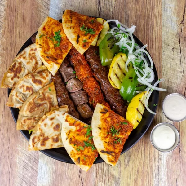 A black plate filled with various kebabs, grilled vegetables, and flatbread, with two dipping sauces.