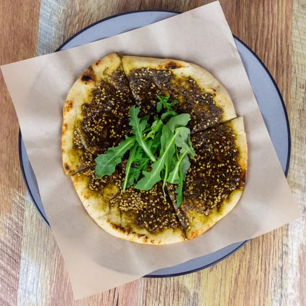 Manakish flatbread with za'atar, sesame seeds, and fresh arugula on a plate with parchment paper.