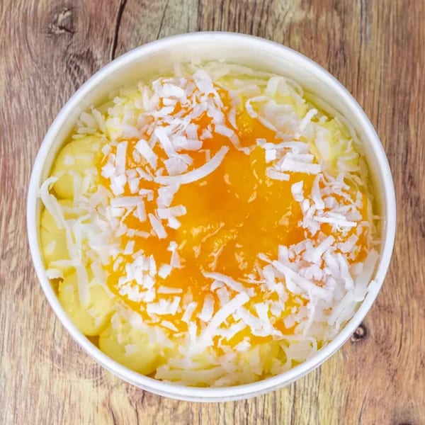 Overhead view of a white bowl filled with yellow dessert, topped with orange jam and shredded coconut.
